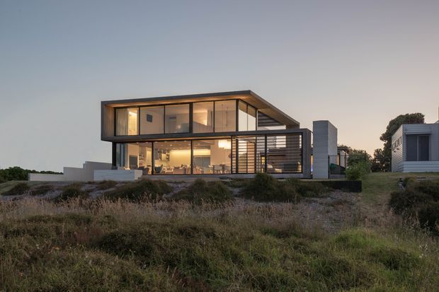 Contemporary two-storey flat-roof house set in a coastal-looking landscaped site at dusk. The home features strong horizontal and rectangular volumes, extensive floor-to-ceiling glazing, dark metal framing, a projecting upper level, and a slatted screened outdoor area. Warm interior lighting is visible through the windows, with a separate small modern outbuilding at right.