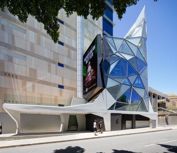 Street-level exterior view of a contemporary urban commercial building with a dramatic angular glass-and-metal geometric facade, projecting canopy, large digital billboard, and adjoining pale stone-clad walls. The faceted blue and clear glazing forms a sculptural crystalline volume against a bright sky.