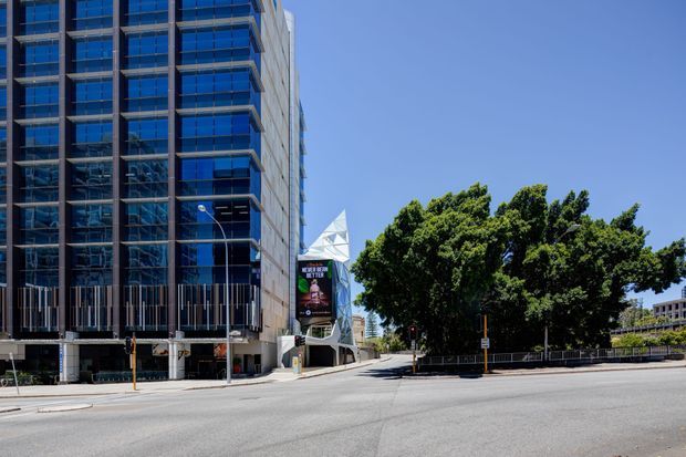 Street-level exterior view of a contemporary multi-storey commercial building with extensive blue reflective glazing, vertical facade fins, projecting balcony-like slabs, and a prominent angular geometric entrance canopy. The building sits beside a broad urban roadway, with mature trees and another building visible in the background under clear daylight.