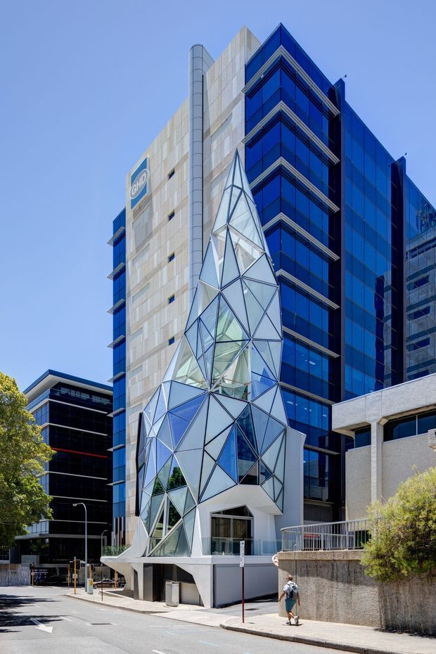 Street-level exterior view of a multi-storey commercial building in an urban setting. The facade combines dark blue curtain-wall glazing with pale stone or concrete panels and a highly distinctive angular, faceted glass-and-metal entrance structure projecting from the lower levels. Adjacent office buildings, a paved street, footpath, signage, and mature street planting are visible.