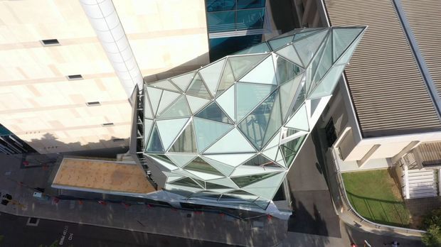 Aerial view of a contemporary urban commercial building featuring a striking faceted glass-and-metal geometric atrium or canopy. The angular glazed structure forms a crystalline roofline between larger rectilinear buildings, with surrounding streets, paving and rooftop elements visible.