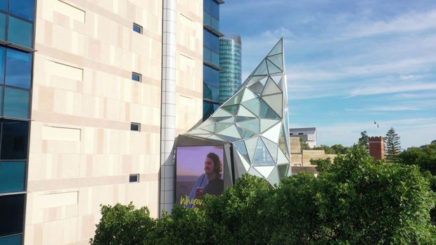 Exterior view of a contemporary multi-storey commercial or institutional building with pale stone or concrete cladding, dark glazed window bands, and a striking angular glass-and-metal geometric facade feature. Mature shrubs partially obscure the lower level, while other urban buildings are visible in the background under a bright sky.