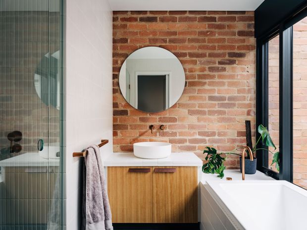 Bright contemporary bathroom with a freestanding white bathtub beside black-framed glazing, a timber floating vanity with white benchtop and round vessel basin, and an exposed red brick feature wall. A round mirror, brushed brass tapware, plants, and a glass shower enclosure complete the space.