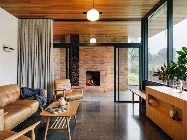Warm mid-century-inspired living room with a timber-lined ceiling, exposed red-brick fireplace wall, polished dark concrete floor, and expansive black-framed glazing. A leather sofa and timber lounge chairs surround a low coffee table, while built-in timber cabinetry runs beneath the window. Full-height curtains and indoor planting add softness to the refined interior.