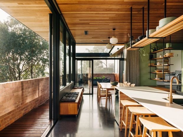 A warm, contemporary open-plan kitchen and dining area with a long white island, timber bar stools, dark green cabinetry, open timber shelving, and a dining table beyond. Full-height black-framed glazing opens onto a timber deck and brick privacy wall, while a richly lined timber ceiling and polished dark floor add warmth and contrast.
