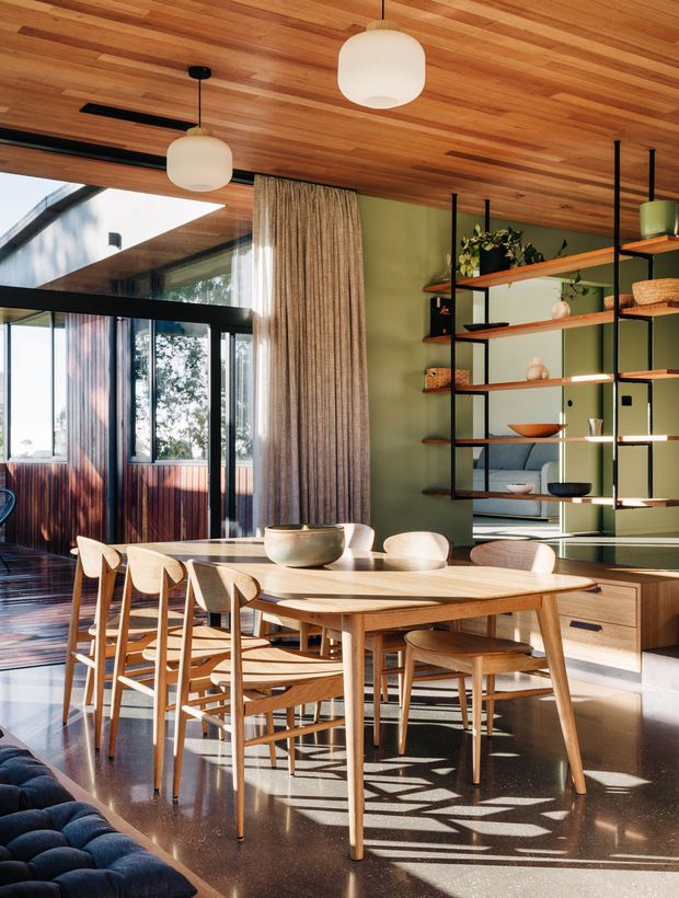 A warm, mid-century-inspired dining room with a large solid timber dining table and matching wooden chairs. The room features a rich timber-lined ceiling, dark polished flooring, olive-green walls, black-framed glazing, sheer curtains, and open black metal shelving with timber shelves and integrated lighting. Strong sunlight enters through the extensive glazing, creating a bright, inviting interior with clear connections to the outdoors.