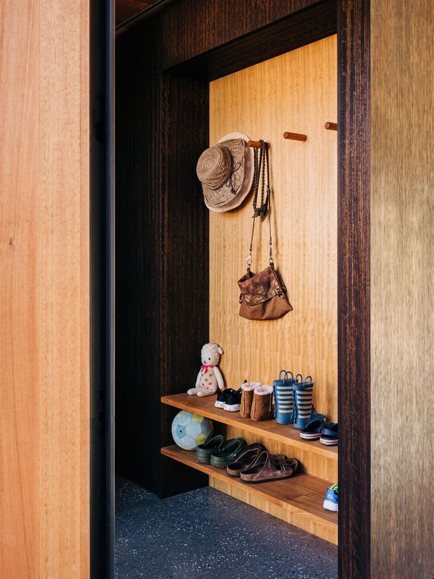 A compact built-in entryway mudroom with dark timber side panels and shelving, a warm light timber back panel with hooks, and open shoe storage. Hats, bags, children's items, boots, shoes, and a toy are arranged on the shelves and hooks. The space has a highly contrasting warm wood palette and a dark speckled floor.