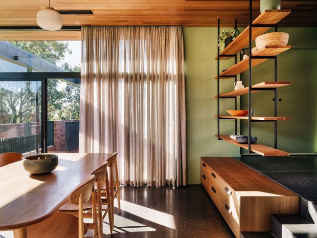 A warm, contemporary dining and kitchen interior with a timber-lined ceiling, large glazed doors opening to an outdoor area, sheer floor-to-ceiling curtains, and a long timber dining table with stools. A dark green wall frames open timber shelving and a built-in timber kitchen cabinet with a dark benchtop. The room has abundant natural light and strong indoor-outdoor connection.