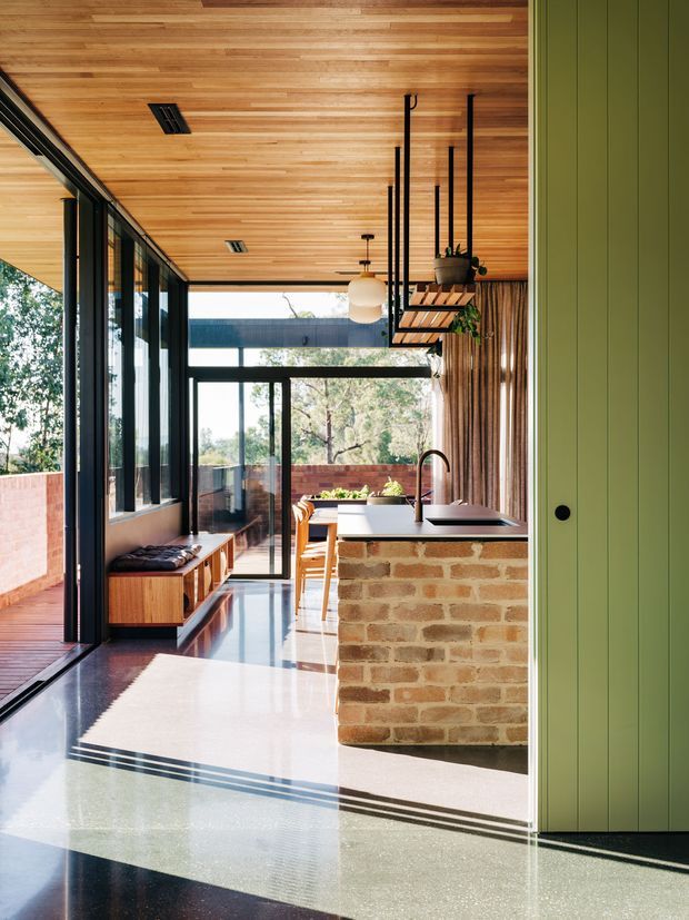 A contemporary open-plan kitchen and dining area with a warm timber-lined ceiling, exposed brick island, dark-framed floor-to-ceiling glazing, and polished concrete or terrazzo flooring. A green vertical-panel door or wall forms the right edge, while a built-in timber bench runs along the glazed side. The space opens toward a timber deck and leafy outdoor setting, with black suspended shelving and pendant lighting adding contrast.
