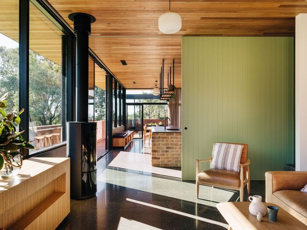 A bright, elongated contemporary living area with floor-to-ceiling black-framed glazing, a timber-lined ceiling, polished dark flooring, and a green vertical-panel feature wall. The space includes a black cylindrical wood-burning stove, timber built-in cabinetry, a brick kitchen island visible beyond, and mid-century-inspired leather and timber seating. Strong natural light creates a warm, refined indoor-outdoor atmosphere.