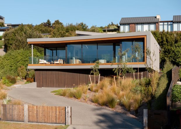 Contemporary two-storey residence on a landscaped sloping site, viewed from the street. The home features a broad low-pitched projecting roof, extensive upper-level glazing, a warm timber-lined soffit and balcony edge, dark vertical timber cladding at the lower level, and an integrated garage. Native-style grasses, shrubs, and timber fencing frame the driveway and facade.