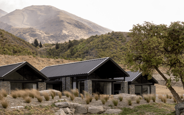 Low-profile contemporary mountain residence composed of several connected gabled volumes, with dark corrugated metal roofs, extensive glazing, and rugged stone-clad columns and walls. The house sits within a dry, naturalistic landscape of tussock grasses and rocks, backed by steep mountainous terrain and native vegetation.
