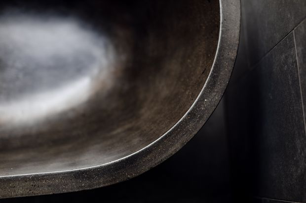 Close-up detail of a dark, round vessel or basin with a heavily textured charcoal-grey rim and reflective interior, set against dark tiled wall surfaces. The image is tightly cropped and primarily highlights the material, finish, and curved form rather than the wider room.