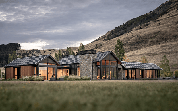 A low, contemporary mountain residence set against a steep, rugged hillside. The building comprises several connected gabled volumes with dark ribbed metal roofs, charcoal window and door frames, warm timber cladding, and a prominent central stone-clad volume. Expansive glazing and warm interior lighting create an inviting appearance, while the blurred grass foreground and mountainous setting emphasize its rural alpine context.