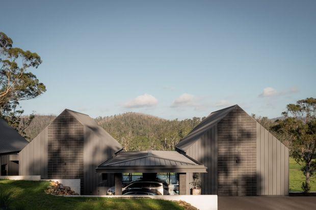 Exterior view of a contemporary multi-volume house in a wooded, semi-rural setting. Several steeply pitched gabled forms are clad in dark vertical and textured metal-like panels, linked by a lower central glazed entry or garage volume with a shallow hipped roof. A driveway and landscaped lawn sit in the foreground, with rolling forested hills beyond.
