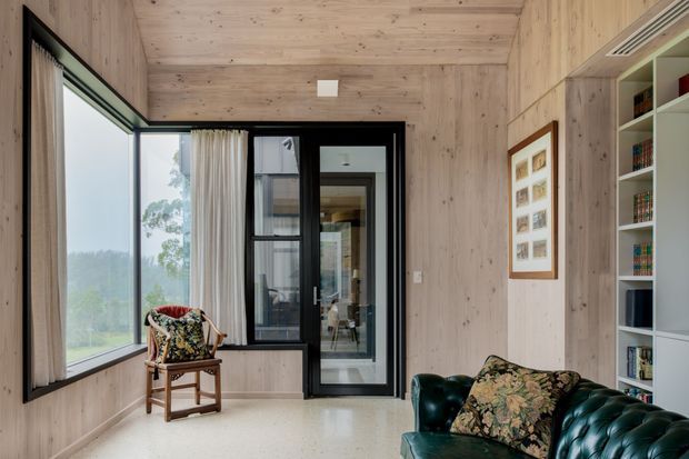 A compact, light-filled living room or sitting area lined with pale timber wall and ceiling panels. Floor-to-ceiling glazing opens toward a leafy landscape, while black-framed internal glazing and a glazed door lead deeper into the home. A dark green leather sofa, patterned cushions, timber armchair, built-in shelving, and framed artwork create a warm, refined interior.