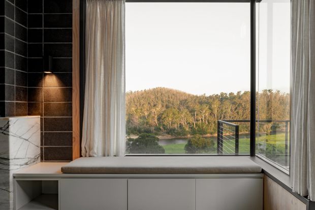 A refined interior window nook with a built-in upholstered bench and white cabinetry beneath large corner glazing. The space features a dark tiled wall with integrated warm lighting, a white marble side surface, timber trim, and sheer curtains. Through the windows is a leafy landscape and water view, while a slim dark metal balustrade is visible outside.