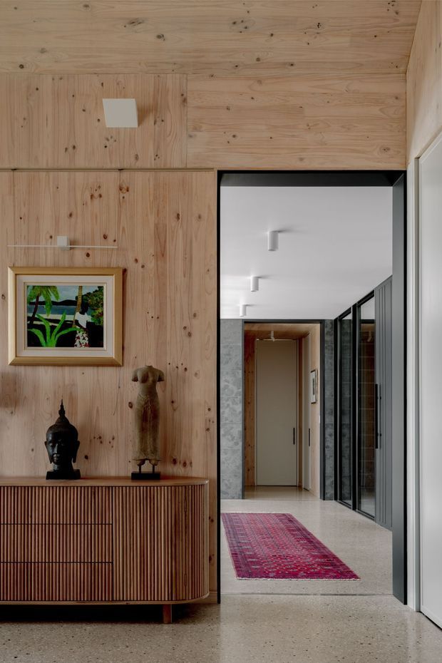 A refined residential entryway or hallway featuring warm natural timber wall panelling, a fluted timber sideboard with rounded corners, framed artwork and sculptural objects. A terrazzo floor continues through a wide opening into a bright corridor with black-framed glazed doors, white walls, and a red patterned runner. The space combines warm wood tones with crisp contemporary detailing and soft, indirect light.