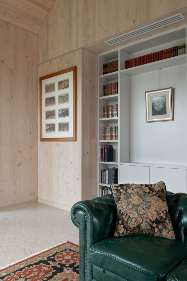 A refined reading or living area with pale timber wall panelling, integrated white built-in bookshelves, framed artwork, and a deep green tufted leather armchair with a floral cushion. A patterned rug and terrazzo-like flooring add texture, while a linear ceiling air vent is integrated above the joinery.