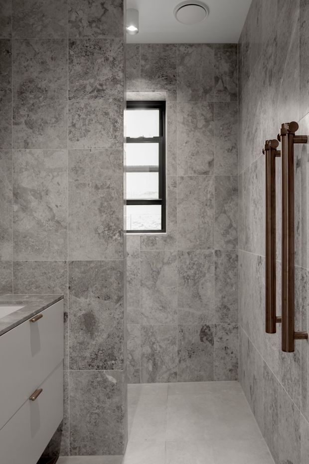A compact contemporary bathroom clad in large-format grey stone-look tiles, with a pale tiled floor, white wall-mounted vanity, narrow black-framed window, recessed ceiling light and circular ventilation grille. A dark bronze heated towel rail is mounted on the right wall.