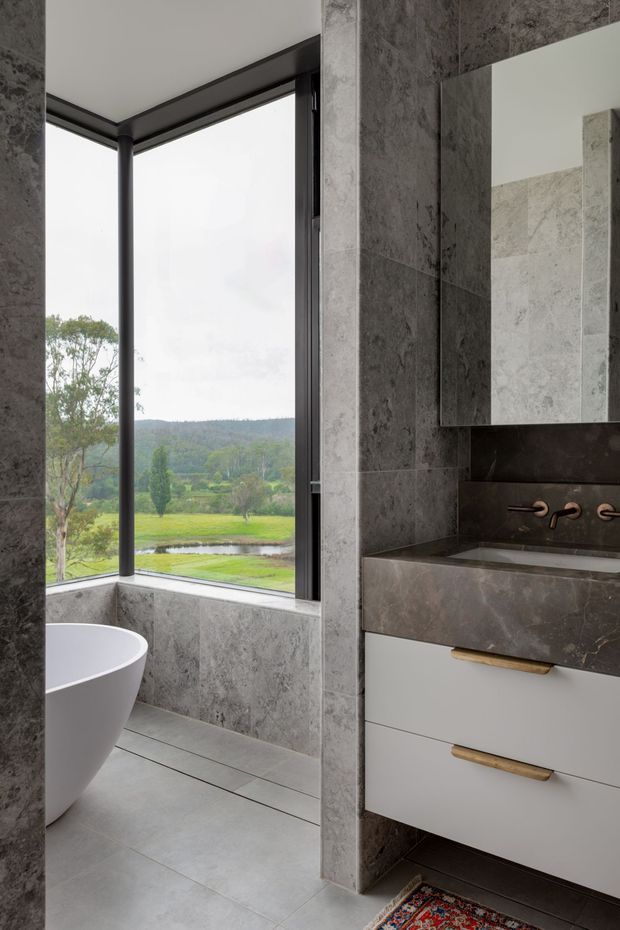 Contemporary bathroom with large grey stone-look wall and floor tiles, a freestanding white bathtub beside expansive black-framed corner glazing, and a floating white vanity with a dark stone benchtop and integrated basin. Copper-toned wall-mounted tapware and timber or brass-toned drawer pulls add warmth. The windows overlook a green rural landscape.