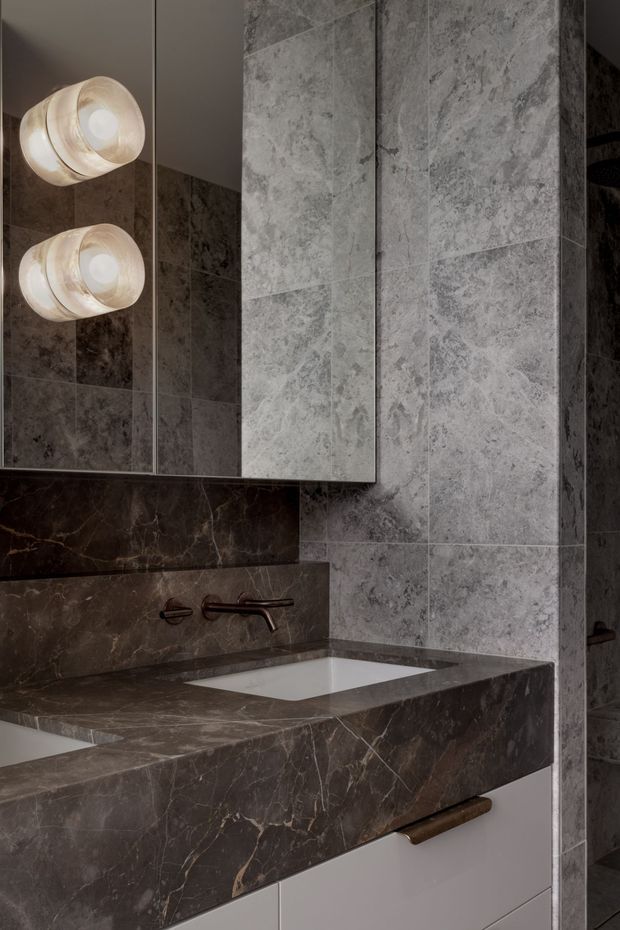 A moody contemporary bathroom vanity featuring a white inset basin, dark brown veined stone benchtop and splashback, grey veined stone wall tiles, pale integrated cabinetry, a wall-mounted dark metal tap, and twin rounded wall lights reflected in the mirrored cabinet.