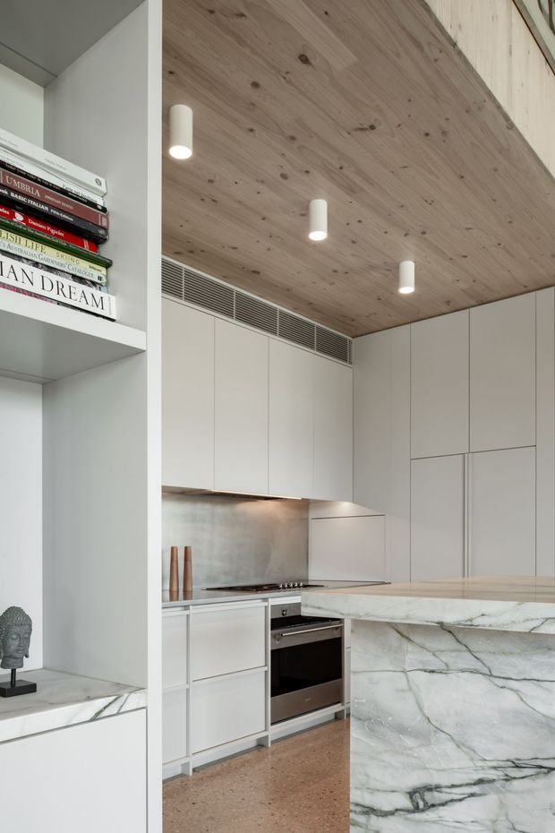 A refined contemporary kitchen with full-height white cabinetry, integrated appliances, a pale veined stone island, and a matching stone-lined shelving unit. The ceiling is clad in light timber boards with recessed linear ventilation and cylindrical pendant-style downlights. A terrazzo-like floor and minimal open shelving with books and a sculptural bust complete the space.