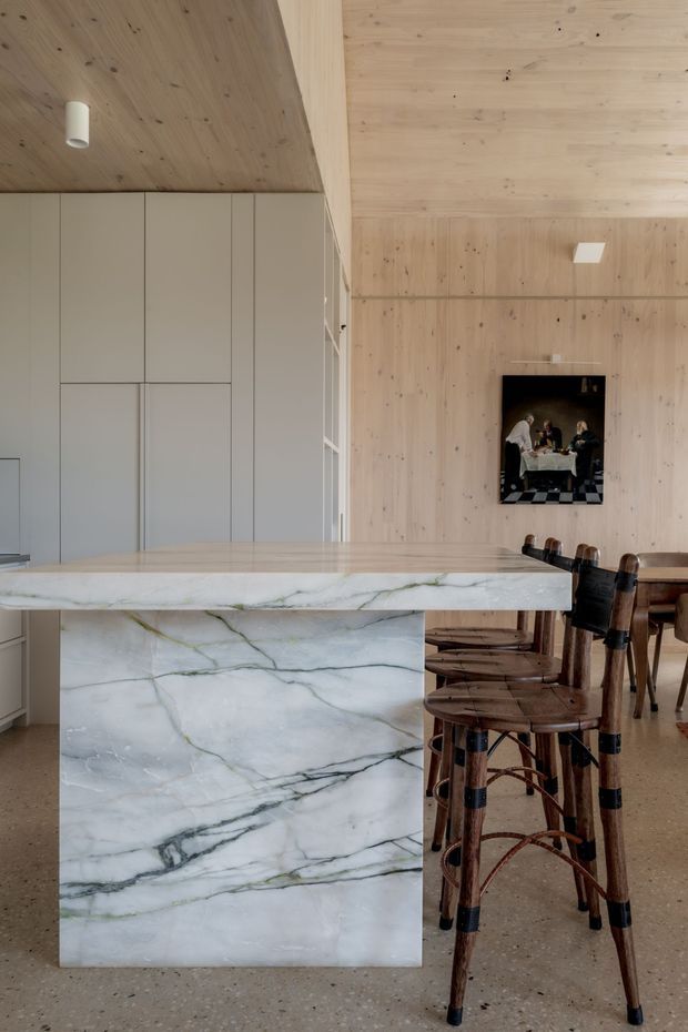 A refined open-plan kitchen and dining area with a large veined natural-stone island, pale timber-lined walls and ceiling, and tall integrated light-grey cabinetry. Woven dark timber bar stools line the island, while a timber dining table and chairs sit behind it. The space has warm natural materials, minimalist detailing, and abundant soft daylight.