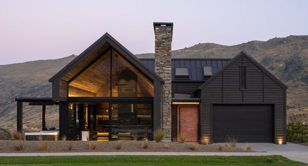 Front exterior view of a contemporary mountain house at dusk, composed of dark horizontal cladding, steep gabled roof forms, extensive floor-to-roof glazing, and a prominent stone chimney. The illuminated timber-lined vaulted interior is visible through the central glazing, while a garage occupies the right wing. Sparse xeriscaped planting and rugged hills form the setting.