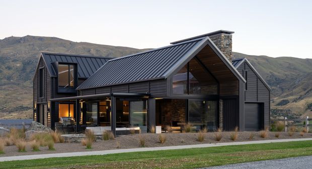 A contemporary multi-level mountain home viewed from the exterior, with dark vertical cladding, a sharply pitched gabled roof, black metal roofing, extensive floor-to-ceiling glazing, and a prominent stone chimney. The house sits in a landscaped, gravelled setting with ornamental grasses and a paved path, backed by rugged hills.