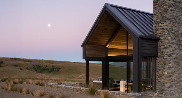 Exterior architectural photograph of a modern alpine or rural pavilion-like house at dusk. The building features a steep gable roof with dark metal roofing, dark timber-clad gable and framing, extensive floor-to-ceiling glazing, and a substantial stacked natural-stone wall. The structure sits in an open tussock-covered landscape with a visible moon and warm interior lighting.