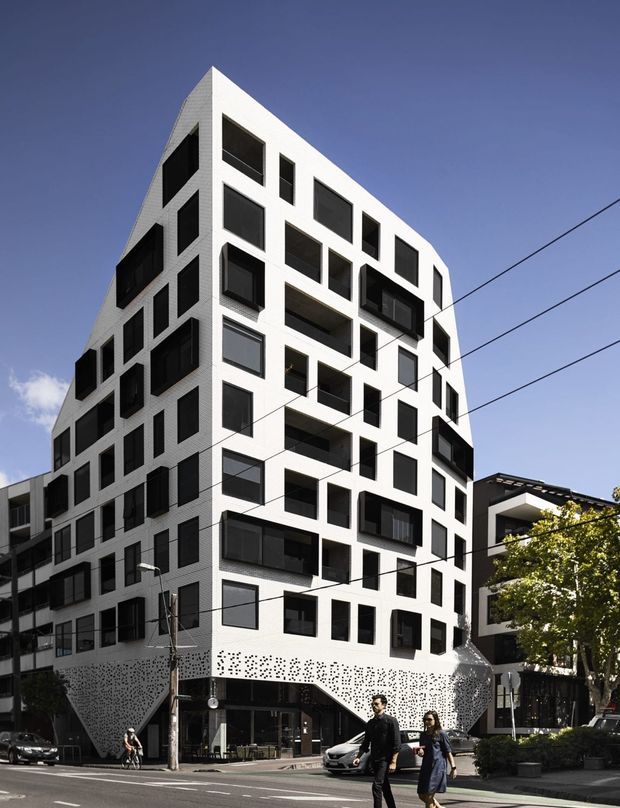 Street-level exterior photograph of a distinctive multi-storey urban apartment or mixed-use building. The white angular facade has a sharply sloping upper edge, numerous irregularly arranged black-framed windows and recessed balconies, and a perforated patterned lower facade. Ground-floor commercial glazing opens onto the sidewalk, with adjacent urban buildings, street infrastructure, trees, and pedestrians visible.