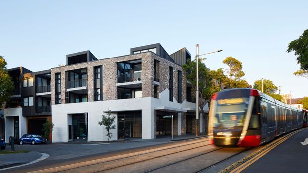 Street-level exterior view of a contemporary multi-storey urban apartment or mixed-use building with a dark brick upper facade, white rendered lower level, balconies, large glazed openings, and varied pitched and flat roof forms. A tram is passing in the foreground along embedded tracks, with mature street trees and neighboring urban buildings visible.