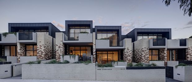 A row of contemporary multi-storey attached townhouses with strong geometric forms, dark metal cladding, pale brick masonry, large glazed openings, projecting balconies, and landscaped front terraces. The street-facing facade is photographed at dusk, showing warm interior lighting and a cohesive modern residential development.