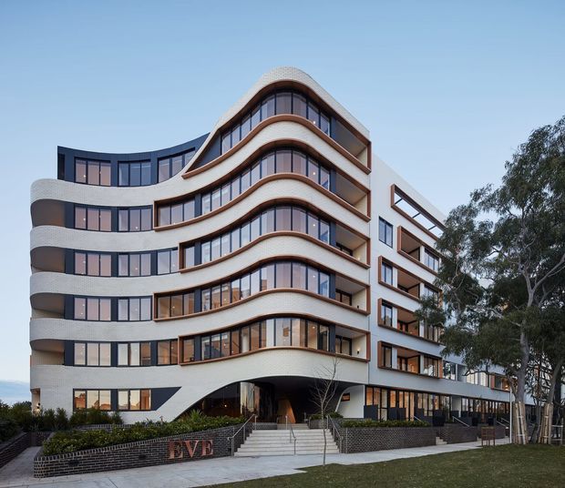 Exterior view of a multi-storey contemporary building with a strongly sculpted facade. Rounded corners, sweeping horizontal balcony and window bands, curved cream-coloured masonry surfaces, dark window frames, and warm timber-toned glazing create a distinctive flowing composition. The building has a recessed arched entrance, landscaped foreground, brick retaining walls, and mature trees beside it.