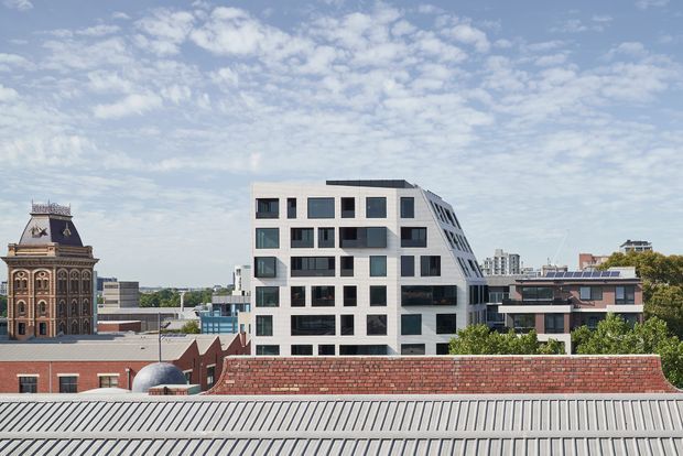 Urban architectural exterior view featuring a contemporary multi-storey apartment or office building with a white geometric facade, dark-framed windows, recessed balconies, and a sloping stepped side elevation. Historic brick and ornate clock-tower architecture appears to the left, with additional low-rise buildings and rooftops in the surrounding cityscape.