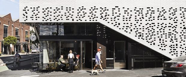 Street-level exterior view of a contemporary urban building with a striking white perforated facade composed of irregular black openings. A sharply sloping facade or roof plane projects over a dark glazed entrance and ground-floor café-style seating, with adjacent brick terrace buildings and street activity visible around it.