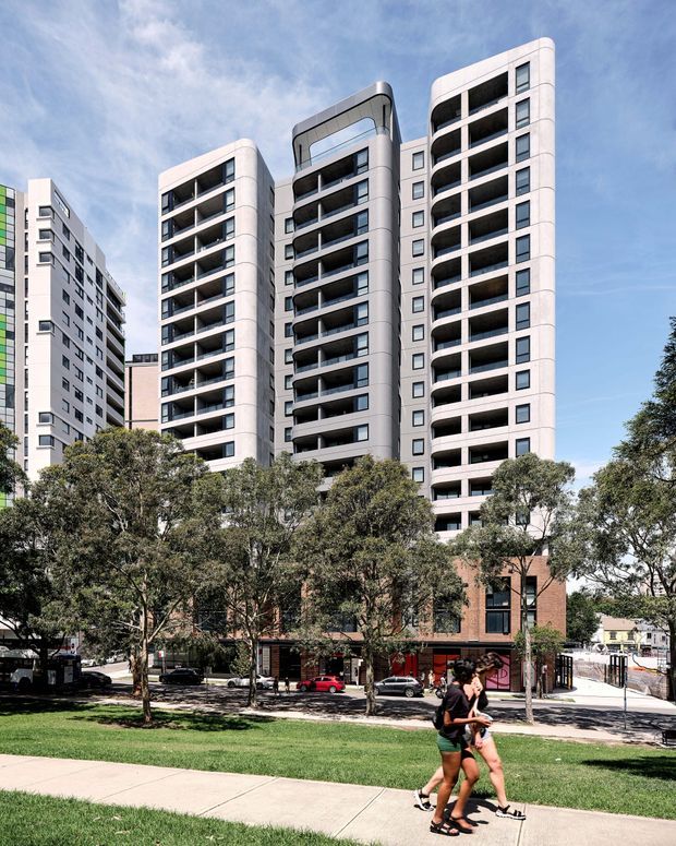 Street-level exterior view of a contemporary multi-storey apartment building, featuring several tall residential towers with rounded corners, recessed balconies, dark glazing, and pale facade panels above a brick-clad podium with ground-floor retail or entry spaces. Mature street trees, a grass verge, sidewalk, and pedestrians occupy the foreground.