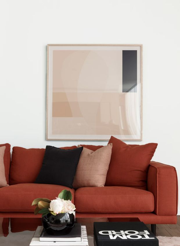 A close-up interior view of a rust-red upholstered sofa against a white wall, styled with rust, blush, and black cushions. A large abstract geometric artwork in a pale timber frame hangs above the sofa. In the foreground, a black vase of pale flowers rests on stacked books and a coffee table. The composition is warm, curated, and contemporary with strong terracotta-red accents.