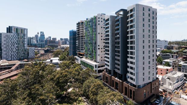 Urban exterior view of a dense cluster of contemporary multi-storey apartment buildings, with prominent high-rise residential towers, balconies, varied facade materials, and a city skyline in the background. Mature trees and lower-rise urban buildings occupy the foreground and surrounding context.
