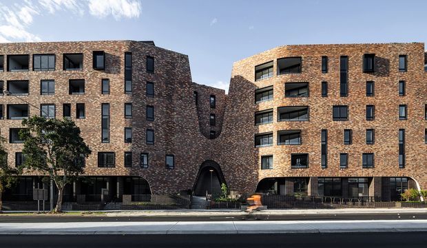 Street-level exterior view of a contemporary multi-storey apartment building composed of two connected brick-clad wings. The facade features repetitive dark-framed windows, recessed balconies, curved corner glazing, a central arched pedestrian entry, and dark ground-floor frontage. Trees, roadway, and sky surround the building.