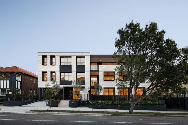 Street-facing photograph of a contemporary multi-residential building, likely a townhouse or apartment development. The facade combines light rendered or masonry panels with dark charcoal sections, extensive black-framed glazing, projecting balconies, recessed terraces, and warm interior lighting. A landscaped frontage with raised planters, small trees, a paved entry path, fencing, and street curb occupies the foreground. Adjacent older residential buildings are visible on either side.