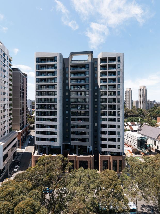 Exterior view of a contemporary multi-storey apartment building in an urban setting. The building has a symmetrical composition with two light concrete-clad wings, a darker recessed central section, extensive balconies and glazed openings, and a brick-clad podium. Surrounding high-rise buildings, streets, mature trees, and neighbouring low-rise buildings are visible under a bright blue sky.