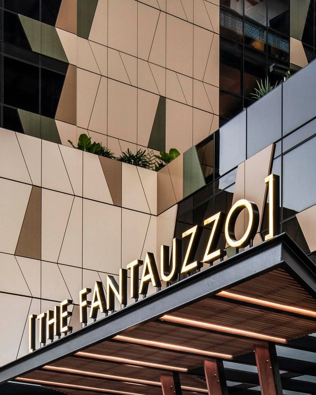 Close-up exterior view of a contemporary multi-storey commercial building entrance, featuring a bold geometric facade of pale beige, black, and muted green panels. Large illuminated signage reads “THE FANTAUZZI,” mounted above a deep entrance canopy with dark metal framing, timber-toned soffit slats, and linear integrated lighting. Planting is incorporated into recessed facade terraces, with glass curtain wall sections visible above and behind.