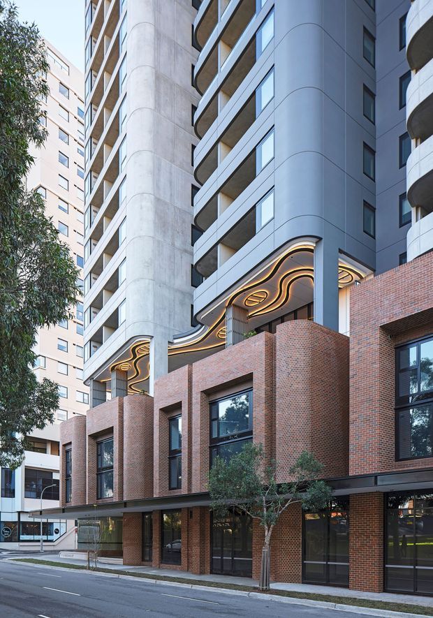 Street-level view of a contemporary multi-storey apartment or mixed-use building in an urban setting. The lower levels feature warm red brick walls, tall black-framed glazing, and projecting brick volumes, while the upper levels have pale concrete or rendered panels, recessed balconies with curved edges, and cylindrical structural elements. A highly distinctive sculptural ceiling canopy with flowing illuminated curves marks the entrance or podium area.