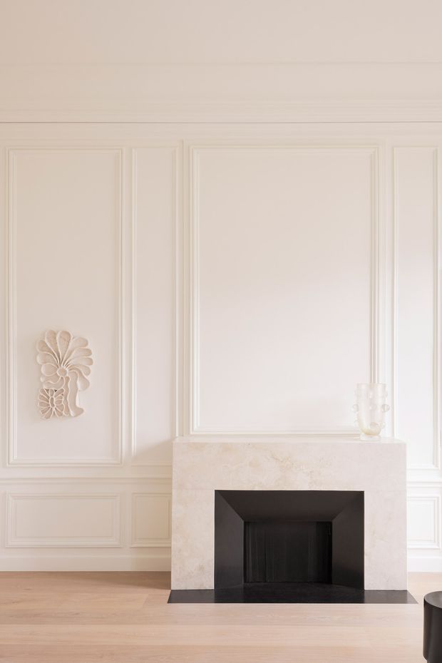 Bright, refined interior detail centred on a sculptural fireplace. The room features pale painted wall panelling with classical moulded rectangular panels, a light stone mantel and surround, black firebox detailing, and light timber flooring. A small decorative wall relief and minimal vessel add subtle ornamentation.