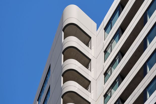 Upward-looking architectural view of a contemporary multi-storey building against a clear blue sky. The facade combines pale smooth cladding, dark recessed balcony undersides, projecting curved balcony edges, and repetitive dark-framed glazing, creating a clean geometric composition.