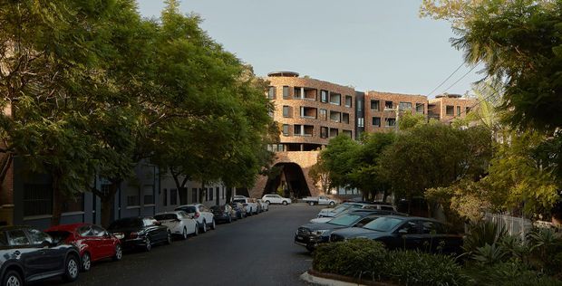 Street-level exterior view of a multi-storey brick apartment or commercial building complex, framed by mature street trees and parked cars. The building has repeated recessed window openings, dark window frames, and a prominent arched vehicle or pedestrian entrance through the central brick mass. The scene appears urban and shaded, with landscaped planting along the street edge.