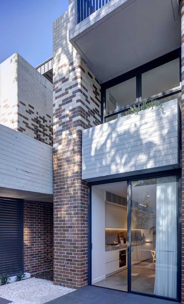 Vertical exterior view of a contemporary multi-storey townhouse or residential building. The facade combines dark red and cream brickwork, pale horizontal cladding panels, dark metal-framed glazing, projecting balconies or overhangs, and a small landscaped entry area. Through the ground-floor glazing, a refined kitchen or living space with light cabinetry, stone surfaces and warm under-cabinet lighting is visible.