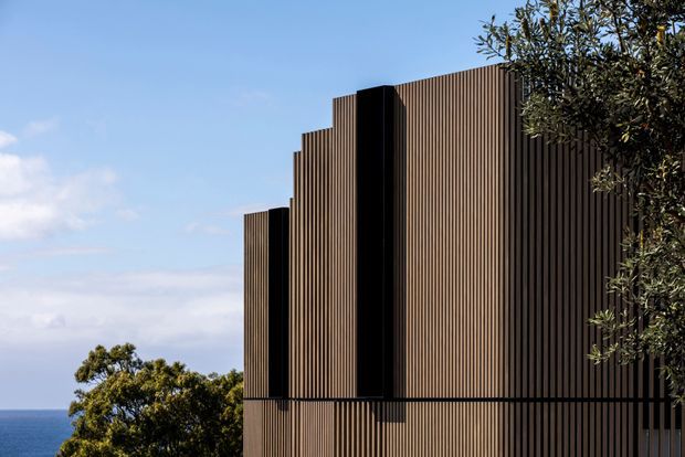 Exterior architectural view of a contemporary building against a coastal horizon. The facade is composed of tall, closely spaced vertical ribbed cladding in a warm dark-brown tone, with stepped volumes and prominent black recessed or projecting vertical elements. Mature trees partially frame the building.